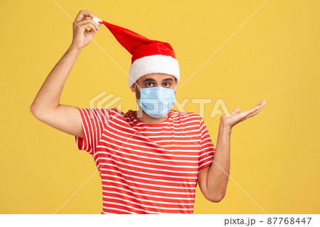 Concerned doubtful man in red t-shirt and santa claus hat with surgical medical mask on face confused with winter holidays under covid-19 quarantine. Indoor studio shot isolated on yellow background Concerned doubtful man in red t-shirt and santa claus hat with surgical medical mask on face confused with winter holidays under covid-19 quarantine. Indoor studio shot isolated on yellow background 87768447