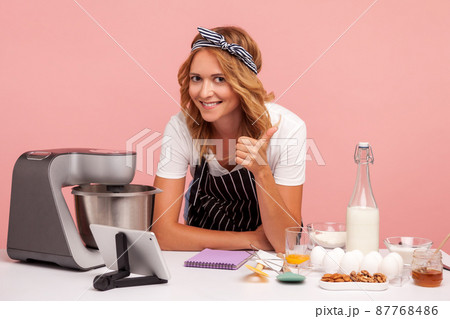 Smiling young adult baker leaning on table full of baking ingredients and food processor, looking at camera and showing thumb up, happy to do pastry. Indoor studio shot isolated on pink background. 87768486