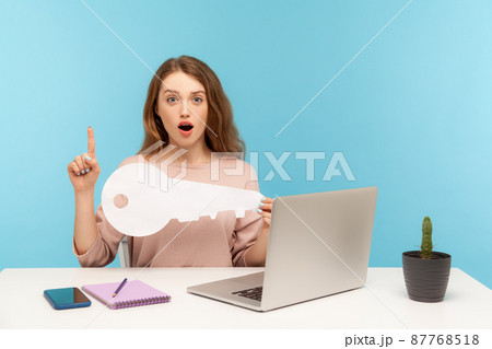 Smart inspired woman employee sitting at workplace with laptop, holding big key and pointing finger up with genius idea, clever solution, found password. indoor studio shot isolated on blue background 87768518
