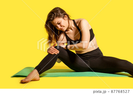 Sportswoman touching painful knee sitting on gym mat, suffering joint injury after yoga, sport workouts, muscle strain, hurting to move sprained ligaments. studio shot isolated on yellow background 87768527