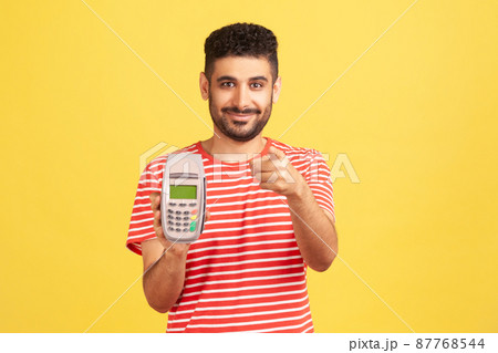 Cheerful bearded man holding in hands pos terminal and pointing finger at camera, suggesting you to use contactless payments. Indoor studio shot isolated on yellow background 87768544