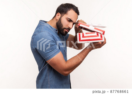 Side view nosy curious man with beard taking a look inside gift box, unpacking present to know what he received, frustrated with content. Indoor studio shot isolated on white background Side view nosy curious man with beard taking a look inside gift box, unpacking present to know what he received, frustrated with content. Indoor studio shot isolated on white background 87768661