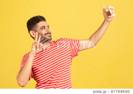 Cheerful popular man blogger in striped t-shirt showing victory gesture looking at smartphone camera, communicating with subscribers, streaming. Indoor studio shot isolated on yellow background Cheerful popular man blogger in striped t-shirt showing victory gesture looking at smartphone camera, communicating with subscribers, streaming. Indoor studio shot isolated on yellow background 87768690