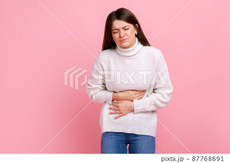 Ill brunette woman suffering acute pain, cramps from indigestion or gastritis, appendicitis symptoms, wearing white casual style sweater. Indoor studio shot isolated on pink background. 87768691