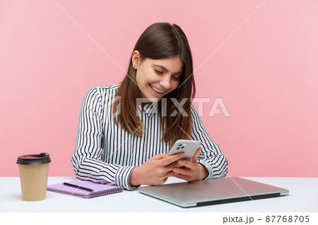 Smiling happy woman office worker in striped shirt chatting or surfing internet at smartphone sitting at workplace, pleasant conversation at messenger. Indoor studio shot isolated on pink background 87768705