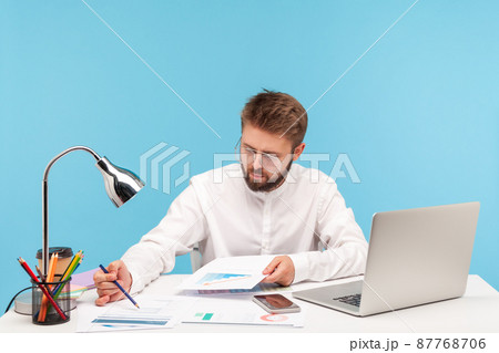 Concentrated business man in white shirt checking and analyzing information looking at graphs and diagrams, sitting at workplace with laptop. Indoor studio shot isolated on blue background Concentrated business man in white shirt checking and analyzing information looking at graphs and diagrams, sitting at workplace with laptop. Indoor studio shot isolated on blue background 87768706