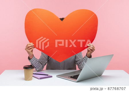 Woman hiding face behind huge red paper heart, using anonymous dating service on laptop, online chat and website. Indoor studio shot isolated on pink background Woman hiding face behind huge red paper heart, using anonymous dating service on laptop, online chat and website. Indoor studio shot isolated on pink background 87768707
