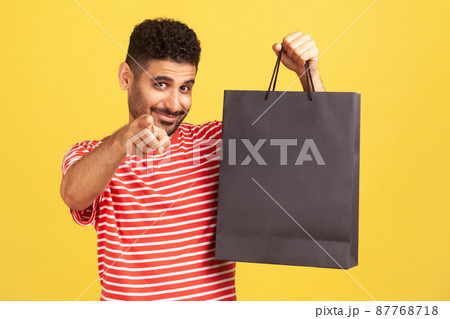 Happy smiling man with beard in striped t-shirt pointing finger at camera holding black paper bag with purchases, inviting you doing shopping. Indoor studio shot isolated on yellow background 87768718
