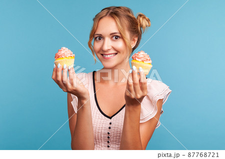 Happy smiling young adult lady in dress with two hair buns showing two tasty cakes, looking at camera with optimism, offers delicious dessert. Indoor studio shot isolated on blue background. Happy smiling young adult lady in dress with two hair buns showing two tasty cakes, looking at camera with optimism, offers delicious dessert. Indoor studio shot isolated on blue background. 87768721