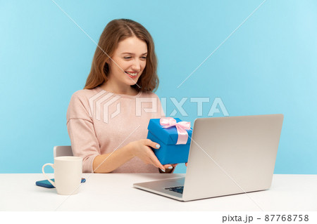 Beautiful woman siting at workplace alone and showing gift box to laptop screen, talking on video call, celebrating birthday alone on self quarantine. indoor studio shot isolated on blue background 87768758