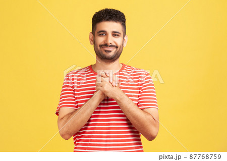 Happy positive man with beard in striped shirt pressing hands together on chest enjoying, calming and comforting himself. Indoor studio shot isolated on yellow background 87768759