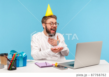 Happy excited man office worker in party cone hat receiving congratulations online holding cake with candle before laptop camera sitting at workplace. Indoor studio shot isolated on blue background 87768762