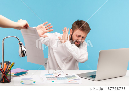 Unhappy bearded man analyst hiding from extra work sitting at workplace with laptop, holding out hands rejecting to sign out dismissal notice. Indoor studio shot isolated on blue background 87768773