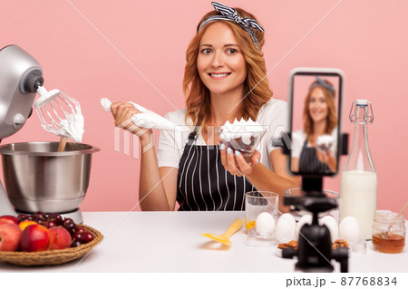 Blonde food blogger making video with tutorial of decoration desserts, looking at smart phone on tripod with smile, holding pastry cone with cream. Indoor studio shot isolated on pink background. Blonde food blogger making video with tutorial of decoration desserts, looking at smart phone on tripod with smile, holding pastry cone with cream. Indoor studio shot isolated on pink background. 87768834