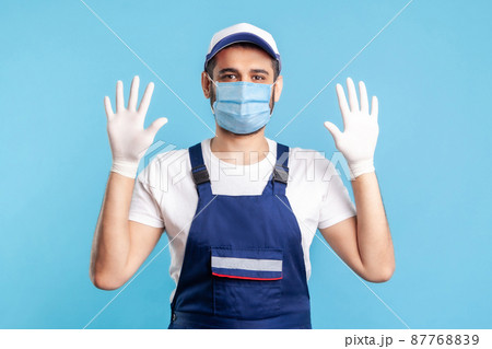 Cheerful handyman in overalls and mask raising hands with surgical gloves, smiling to camera. Safety clothes, protection and hygiene in service industry, courier delivery, housekeeping maintenance. 87768839
