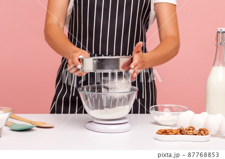 Cropped image of female confectioner sifts flour for doing homemade pastry, lady wearing apron, holds sieve in hands, surrounded with bakery ingredients. Indoor studio shot isolated on pink background 87768853