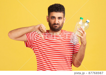 Sad unhappy bearded man showing thumbs down gesture holding in hand plastic bottles, dissatisfied with environmental pollution, worrying about ecology. Indoor studio shot isolated on yellow background 87768987