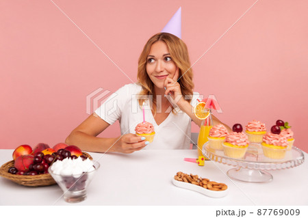 Pensive dreamy woman with blonde hair holding cake with candle and looking away with thoughtful expression, thinking about birthday desire. Indoor studio shot isolated on pink background. Pensive dreamy woman with blonde hair holding cake with candle and looking away with thoughtful expression, thinking about birthday desire. Indoor studio shot isolated on pink background. 87769009