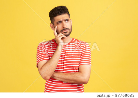 Doubtful bearded man in striped t-shirt contemplating leaning head on hand, pondering difficult question, trying to make right choice. Indoor studio shot isolated on yellow background Doubtful bearded man in striped t-shirt contemplating leaning head on hand, pondering difficult question, trying to make right choice. Indoor studio shot isolated on yellow background 87769035