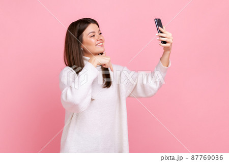 Positive brunette woman taking selfie or having livestream, pointing finger down, subscribe her vlog, wearing white casual style sweater. Indoor studio shot isolated on pink background. Positive brunette woman taking selfie or having livestream, pointing finger down, subscribe her vlog, wearing white casual style sweater. Indoor studio shot isolated on pink background. 87769036