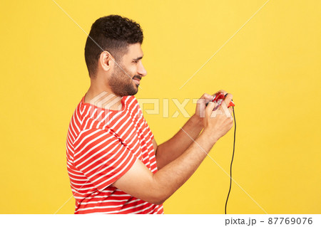 Profile portrait bearded man in red striped t-shirt holding in hands red gamepad joystick, grimacing playing video games. Indoor studio shot isolated on yellow background 87769076