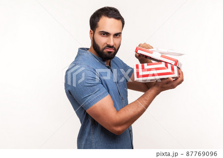 Side view unsatisfied displeased man holding in hands unpacked gift box, frustrated with content, bad present. Indoor studio shot isolated on white background Side view unsatisfied displeased man holding in hands unpacked gift box, frustrated with content, bad present. Indoor studio shot isolated on white background 87769096