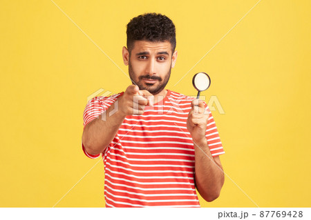 Serious confident man private detective in striped t-shirt holding in hand magnifying glass and pointing finger on you looking at camera, spying. Indoor studio shot isolated on yellow background Serious confident man private detective in striped t-shirt holding in hand magnifying glass and pointing finger on you looking at camera, spying. Indoor studio shot isolated on yellow background 87769428