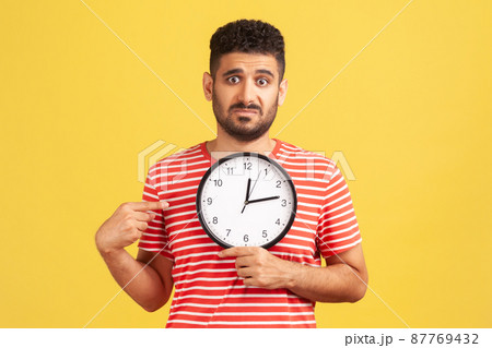 Look at time. Unhappy impatient bearded guy in striped t-shirt pointing big clock and looking displeased irritated, showing wall watches to hurry up. Indoor studio shot isolated on yellow background 87769432