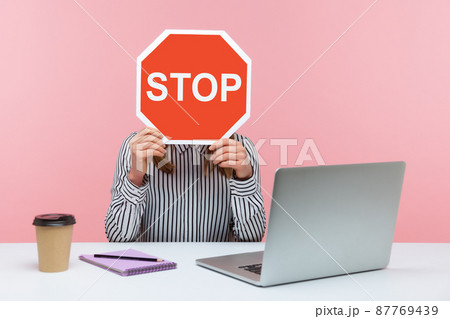 Female office worker in striped shirt sitting at workplace hiding face behind red stop traffic sign avoiding conflicts, afraid of workplace bullying. Indoor studio shot isolated on pink background Female office worker in striped shirt sitting at workplace hiding face behind red stop traffic sign avoiding conflicts, afraid of workplace bullying. Indoor studio shot isolated on pink background 87769439