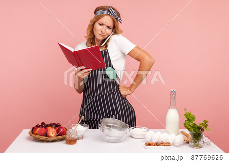 Puzzled young adult woman baker taking phone and asking help with preparing pastry, holding notebook in hands, surrounded with bakery ingredients. Indoor studio shot isolated on pink background. Puzzled young adult woman baker taking phone and asking help with preparing pastry, holding notebook in hands, surrounded with bakery ingredients. Indoor studio shot isolated on pink background. 87769626