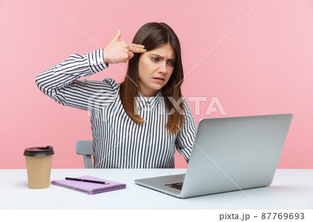 Depressed woman office worker holding finger gun near head talking video call on laptop, expressing extreme fatigue and boredom from job. Indoor studio shot isolated on pink background 87769693