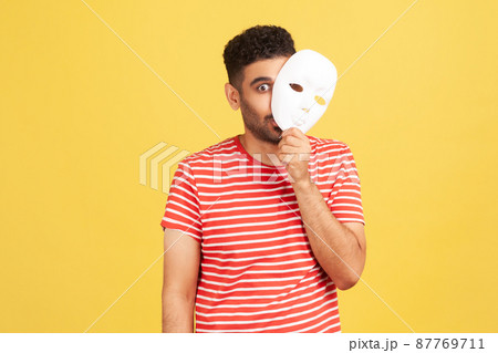Funny crazy man with beard in striped t-shirt peeping out white face mask, hiding his real feelings, pretending to be another person. Indoor studio shot isolated on yellow background 87769711