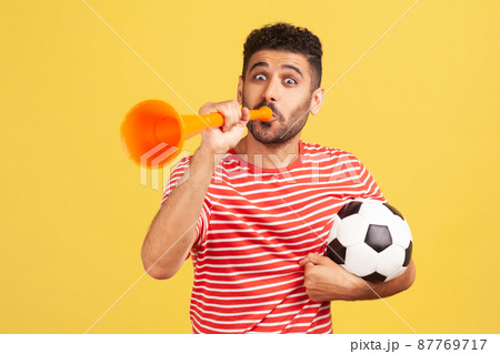 Very happy cheerful man with beard in striped t-shirt blowing in big bullhorn holding football ball, celebrating championship beginning. Indoor studio shot isolated on yellow background Very happy cheerful man with beard in striped t-shirt blowing in big bullhorn holding football ball, celebrating championship beginning. Indoor studio shot isolated on yellow background 87769717