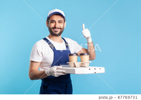 Delivery service. Friendly optimistic courier in overalls holding coffee and pizza box, wearing safety gloves offering drinks food and showing thumbs up, like gesture. indoor studio shot, isolated 87769731