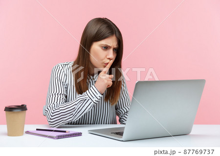 Please, keep secret. Concerned woman employee sitting at workplace shushing with silence gesture, talking on video call, online communication via laptop. Indoor studio shot isolated on pink background 87769737