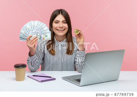 Positive woman office worker holding and showing fan of hundred dollar banknotes and bitcoin crypto coin looking at camera with toothy smile. Indoor studio shot isolated on pink background Positive woman office worker holding and showing fan of hundred dollar banknotes and bitcoin crypto coin looking at camera with toothy smile. Indoor studio shot isolated on pink background 87769739