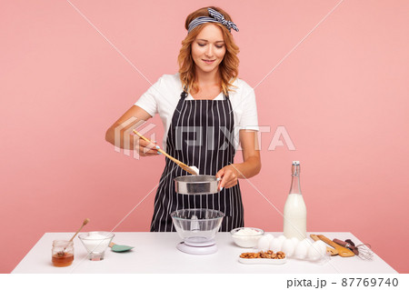 Good looking concentrated female baker preparing pastry, adding flour to sieve, doing pastry, wearing white T-shirt and striped apron. Indoor studio shot isolated on pink background. Good looking concentrated female baker preparing pastry, adding flour to sieve, doing pastry, wearing white T-shirt and striped apron. Indoor studio shot isolated on pink background. 87769740
