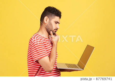 Profile portrait thoughtful pensive man with beard in striped t-shirt holding laptop and rubbing chin looking at display, thinking over new business project. Indoor isolated on yellow background 87769741
