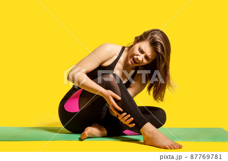 Sportswoman shouting from pain in leg, sitting on gym mat touching injured caviar, suffering calf muscle strain after yoga workout, hurting to move sprained ligaments. indoor studio shot, isolated 87769781