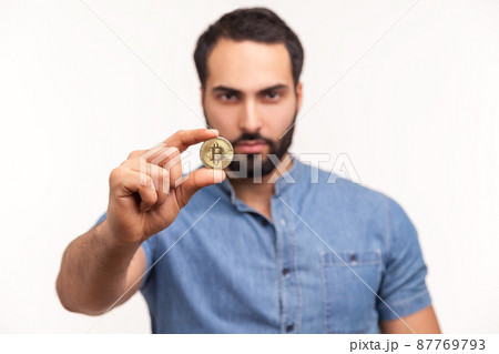 Serious bearded man holding in fingers and showing golden coin bitcoin crypto currency, electronic virtual money. Indoor studio shot isolated on white background 87769793