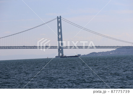 青い海面の海とタンカーと明石海峡大橋と淡路島と雲と青空と霞む山の見える風景 87775739