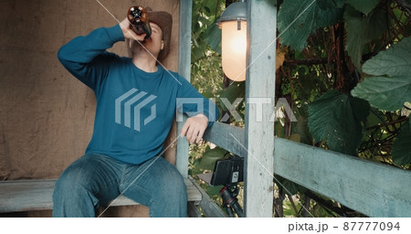 Man in a cowboy hat sits on the porch of a village house and communicates online by phone. He drinks beer from a bottle and rejoices. The concept of remote communication during lockdowns and pandemics 87777094