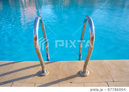Close up of swimming pool stainless steel handrail descending into tortoise clear pool water. Accessibility of recreational activities concept. Close up of swimming pool stainless steel handrail descending into tortoise clear pool water. Accessibility of recreational activities concept. 87786004