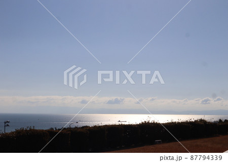 逆光で暗い丘から青空と雲とキラキラ光る水面と青い海面の海と船と霞む山と街の対岸の見える風景 87794339