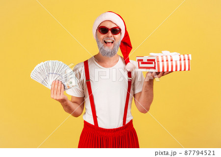 Cheerful positive elderly man in sunglasses and santa claus hat holding fan of dollars and gift box smiling at camera, preparing presents on holidays. Indoor studio shot isolated on yellow background Cheerful positive elderly man in sunglasses and santa claus hat holding fan of dollars and gift box smiling at camera, preparing presents on holidays. Indoor studio shot isolated on yellow background 87794521