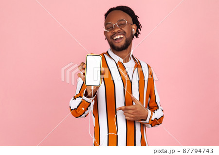 Cheerful positive man with dreadlocks and earphones in striped stylish shirt pointing finger at white screen of smartphone, advertising app. Indoor studio shot isolated on pink background Cheerful positive man with dreadlocks and earphones in striped stylish shirt pointing finger at white screen of smartphone, advertising app. Indoor studio shot isolated on pink background 87794743