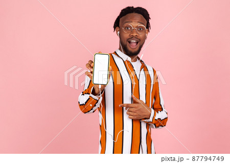 Surprised confused man with dreadlocks and earphones in trendy striped shirt pointing finger at smartphone with white screen, advertisement. Indoor studio shot isolated on pink background Surprised confused man with dreadlocks and earphones in trendy striped shirt pointing finger at smartphone with white screen, advertisement. Indoor studio shot isolated on pink background 87794749