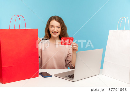 Happy woman buyer surrounded by packages smiling and holding credit card with Sale inscription, making purchases online on laptop using shopping loan. indoor studio shot isolated on blue background 87794848
