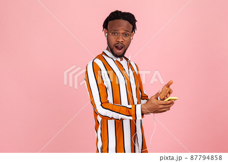Surprised bearded african man with dreadlocks in striped shirt listening to music on smartphone in earphones and looking at camera with astonishment. Indoor studio shot isolated on pink background 87794858