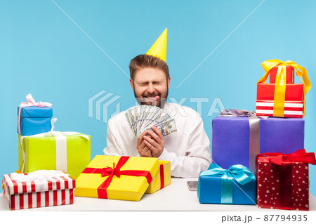 Happy excited bearded man in party hat sincerely rejoicing showing tongue and holding dollar cash, unboxing present, receiving cash bonus. Indoor studio shot isolated on blue background 87794935